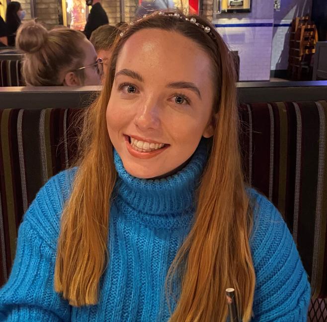 Young lady with long brown hair, wearing a blue jumper and smiling at the camera.