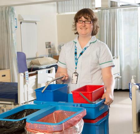 Female student in NHS staff uniform, holding buckets and cleaning equipment in hospital ward.